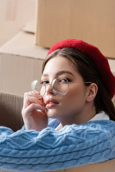 Portrait of trendy woman in beret and eyeglasses looking at camera near cardboard boxes — Stockfoto