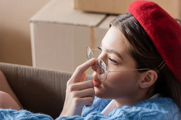 Young woman in beret touching eyeglasses near blurred cardboard boxes — Photo de stock