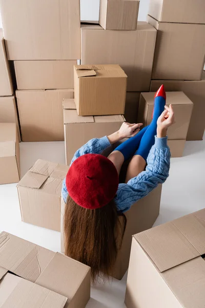 Back view of brunette woman in beret and knee socks sitting in package near cardboard boxes on white background — Stock Photo