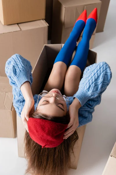 High angle view of stylish woman in eyeglasses touching beret while sitting in package near cardboard boxes on white background - foto de stock