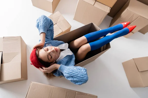 Top view of stylish woman in beret and eyeglasses sitting in package near cardboard boxes on white background — Foto stock