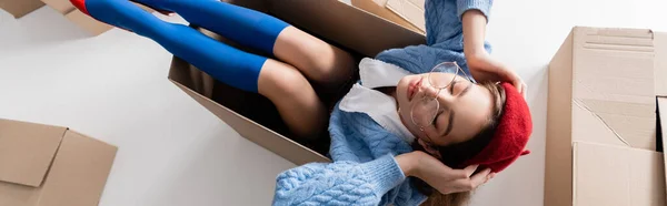 Top view of woman in eyeglasses holding beret while sitting in package on white, banner — Stock Photo