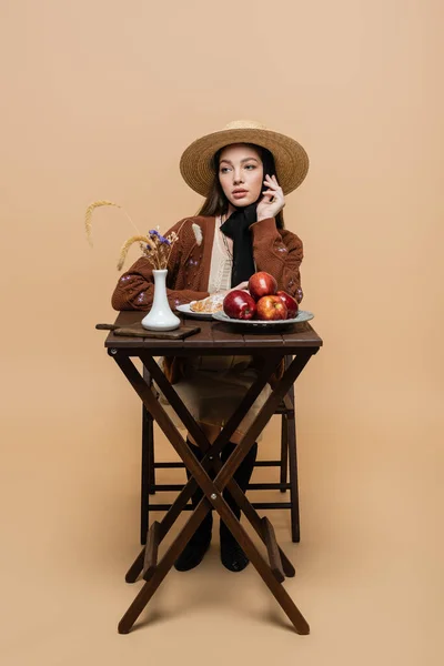 Модная молодая женщина в винтажной одежде сидит рядом с едой и растениями на столе на бежевом — стоковое фото
