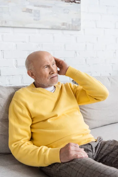 Elderly man touching head while sitting on couch at home — Stock Photo