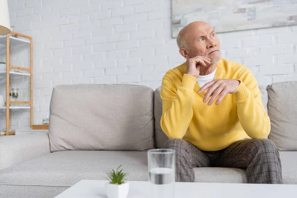 Elderly man sitting near blurred glass of water in living room at home — Stock Photo