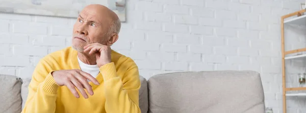 Elderly man looking away while sitting on couch at home, banner — Stockfoto