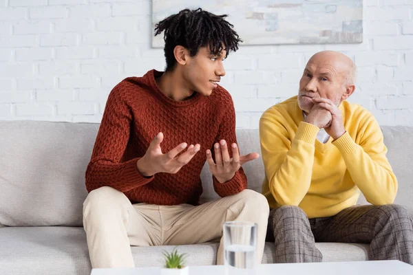 Smiling african american grandson talking to grandparent on couch in living room - foto de stock