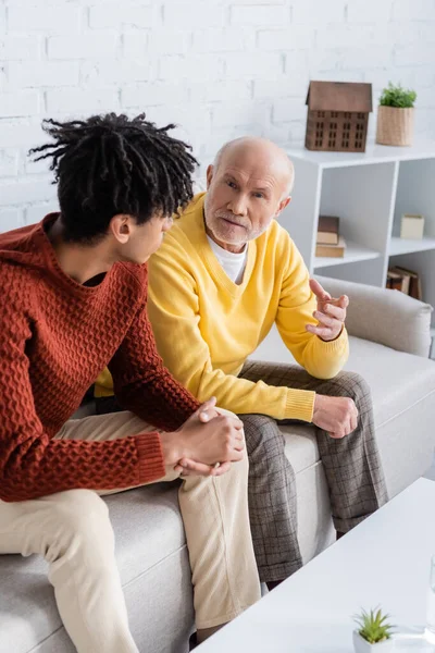 Старший мужчина разговаривает с африканским внуком на диване дома — стоковое фото