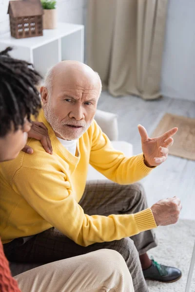 Недовольный пожилой человек, разговаривающий дома с размытым африканским внуком — стоковое фото