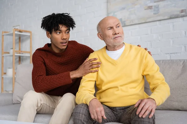 African american grandson hugging disappointed grandpa on couch at home - foto de stock