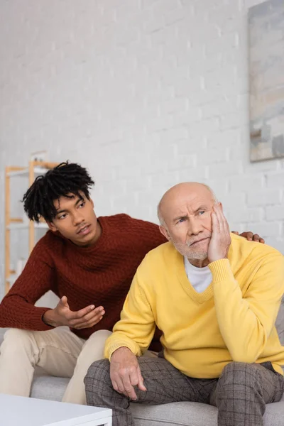 Upset elderly man sitting near african american grandson talking at home — Photo de stock