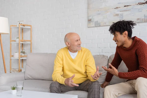 Веселый межрасовый дедушка и внук разговаривают дома на диване — стоковое фото