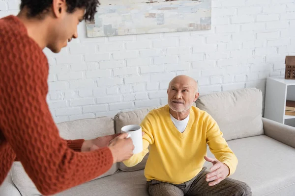 Blurred african american grandson giving cup of tea to smiling granddad at home — Foto stock