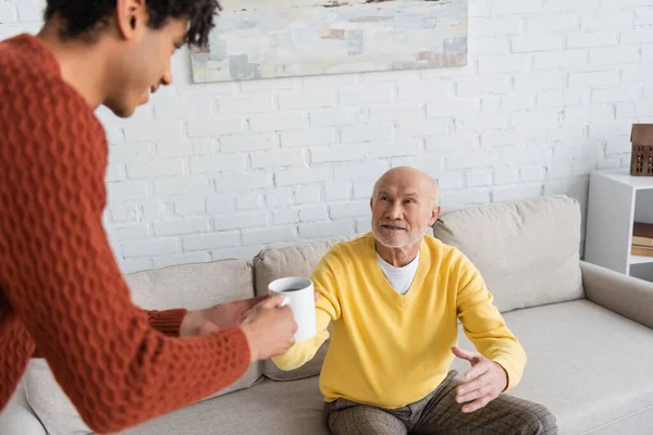 Blurred african american grandson giving cup of tea to smiling granddad at home - foto de stock