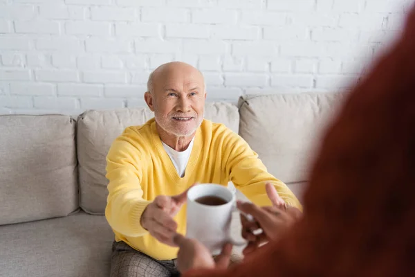 Blurred african american man holding cup of tea near smiling granddad on couch — Stockfoto