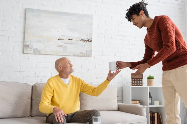 Side view of african american grandson giving cup to smiling grandparent at home — Stock Photo