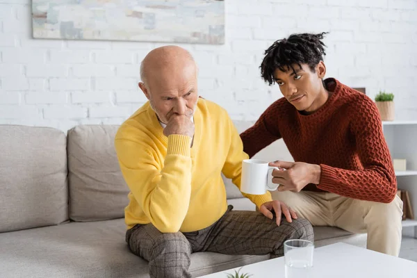 African american grandson holding cup near upset grandparent at home - foto de stock