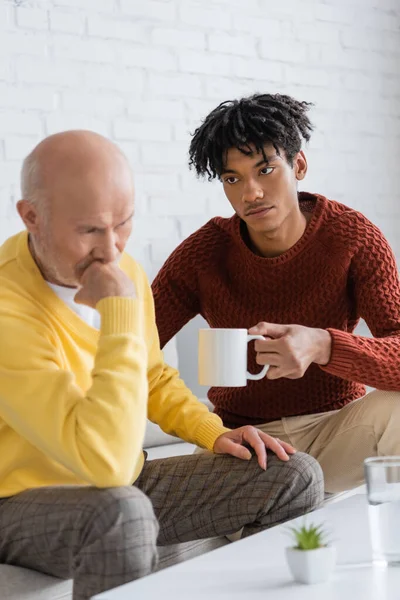 Young african amrican man holding cup near displeased granddad at home — Stock Photo