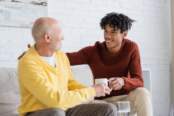 Positive african american grandson giving cup to grandpa in living room — Photo de stock