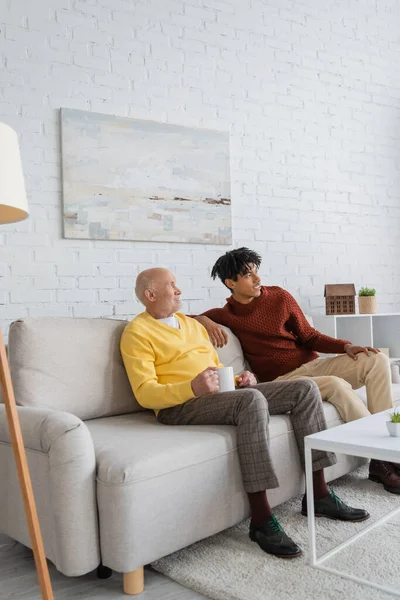 Smiling african american man sitting near granddad with cup on couch at home — Fotografia de Stock