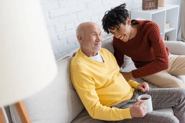 Happy african american grandson sitting near granddad with cup on couch at home - foto de stock