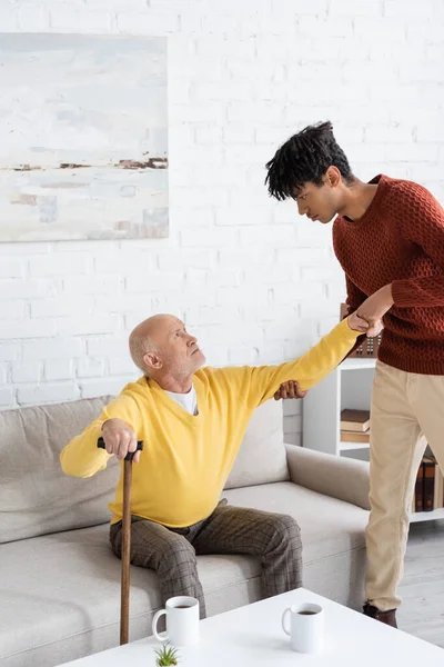 African american grandson helping granddad with walking cane in living room - foto de stock