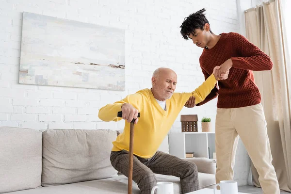 African american grandson helping displeased granddad with walking cane at home — Fotografia de Stock