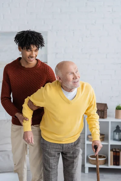 Smiling african american grandson helping grandpa with walking cane at home - foto de stock