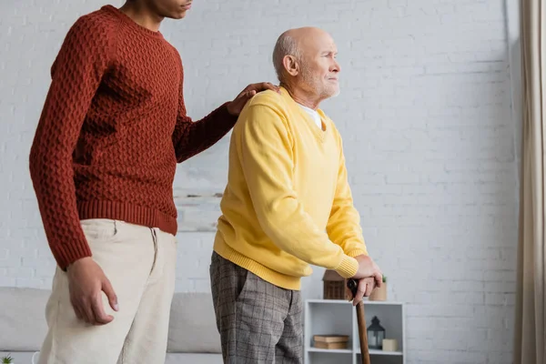 African american grandson hugging sad grandpa with walking cane at home — Stockfoto