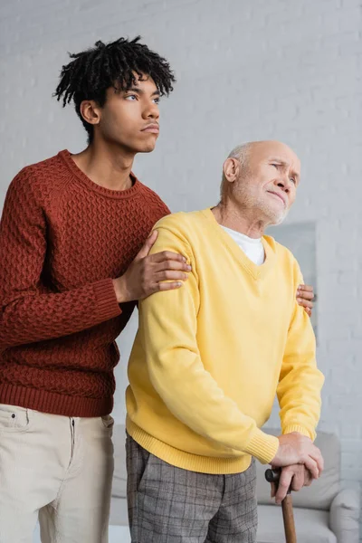 Young african american man hugging upset grandparent with walking cane at home - foto de stock