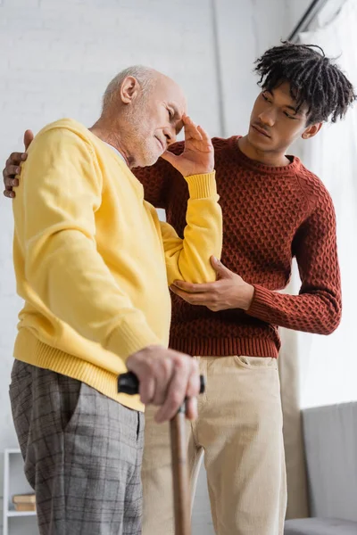 Low angle view of displeased pensioner holding walking cane near african american grandson at home - foto de stock