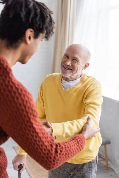 Cheerful pensioner with walking cane holding hand of blurred african american grandson at home — Photo de stock