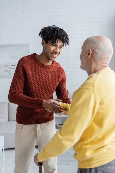 Positive african american grandson holding hand of grandparent with walking cane in living room - foto de stock