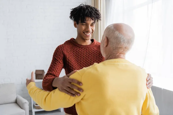 Smiling african american man hugging and looking at blurred granddad at home - foto de stock