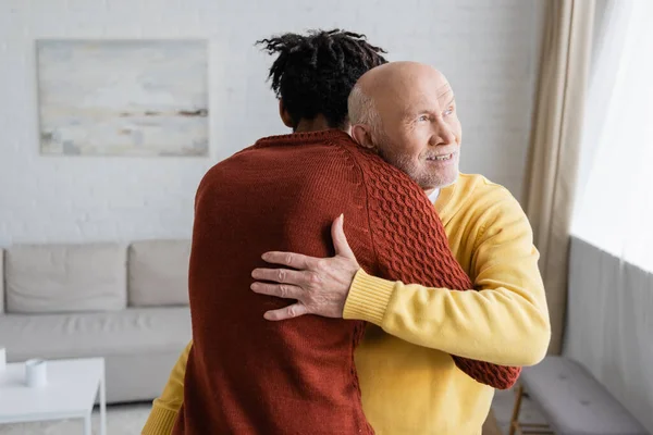 Cheerful senior man hugging african american grandson in living room - foto de stock