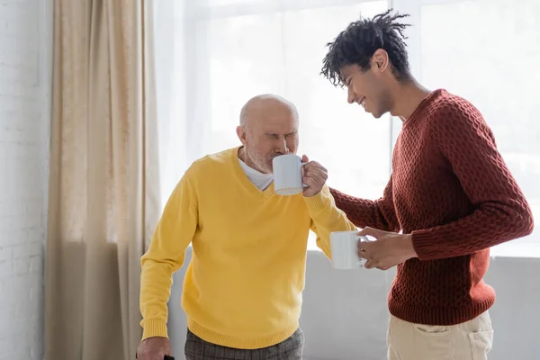 Smiling african american man standing near granddad drinking tea at home — Foto stock