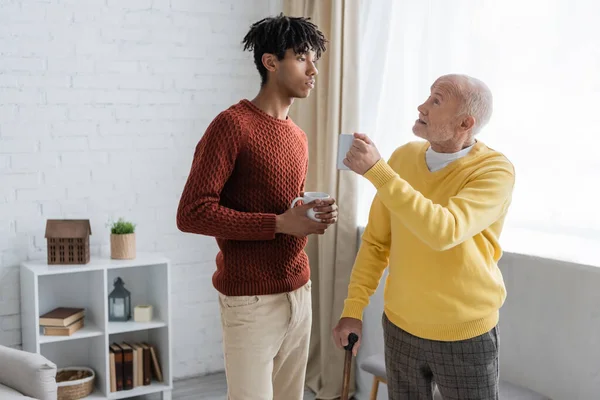 Senior man holding walking cane and cup while talking to african american grandson at home — Fotografia de Stock