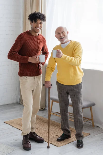 Cheerful interracial granddad and grandson holding cups and looking at camera at home — Fotografia de Stock