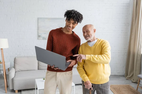 Senior man with walking cane pointing with finger near african american grandson with laptop at home — Stockfoto