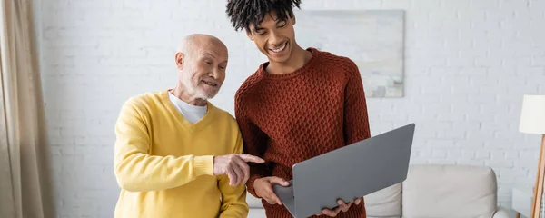 Позитивный пенсионер указывает на ноутбук рядом с африканским внуком Америки дома, баннер — стоковое фото