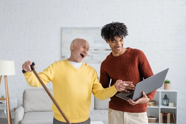 Excited pensioner with walking cane pointing at laptop near african american grandson at home - foto de stock