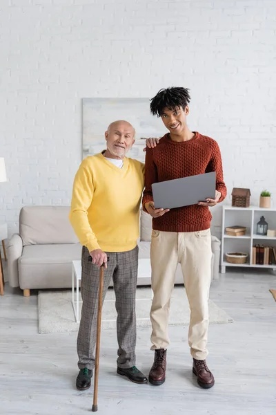 Smiling pensioner with walking cane looking at camera near african american grandson with laptop at home — Stockfoto