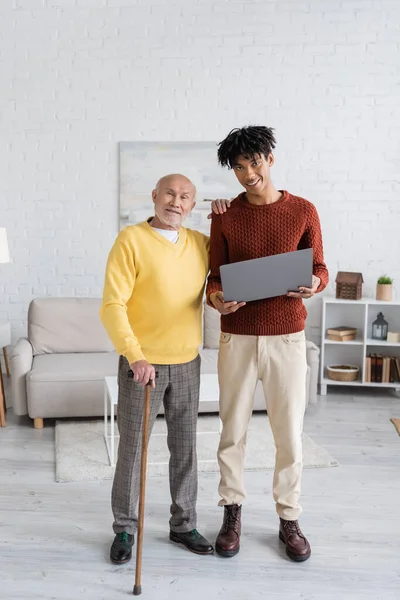 Smiling pensioner with walking cane looking at camera near african american grandson with laptop at home — Fotografia de Stock