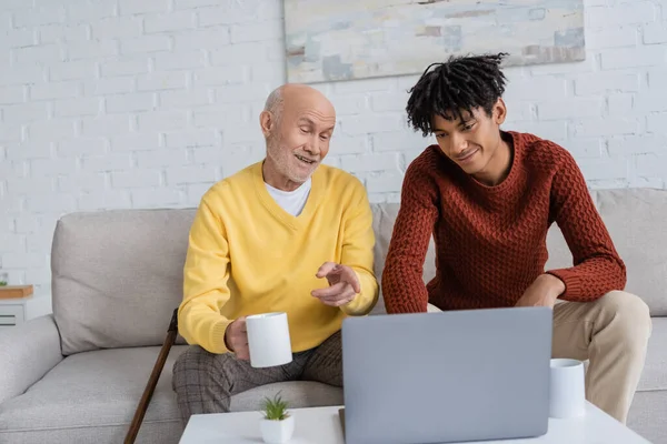 Cheerful pensioner pointing at laptop and holding cup near african american grandson at home — Stockfoto