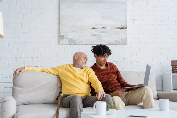 Granddad and african american grandson using laptop near walking cane and cups in living room — Foto stock