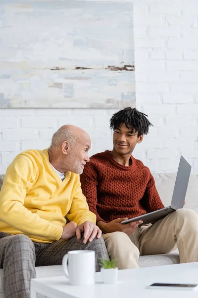 Senior man talking near smiling african american grandson with laptop at home — Photo de stock