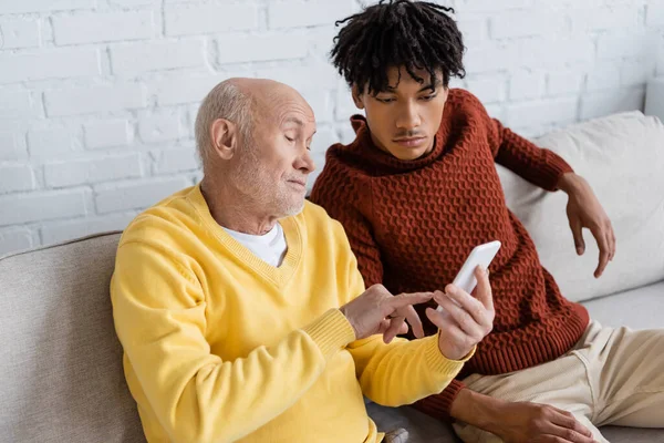 Літній чоловік вказує на смартфон біля афроамериканського онука на дивані вдома — стокове фото