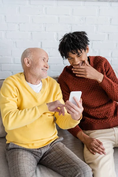 Cheerful pensioner holding smartphone near african american grandson on couch — Stock Photo