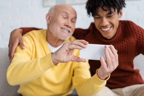 Smartphone in hands of blurred pensioner near african american grandson at home — Stock Photo