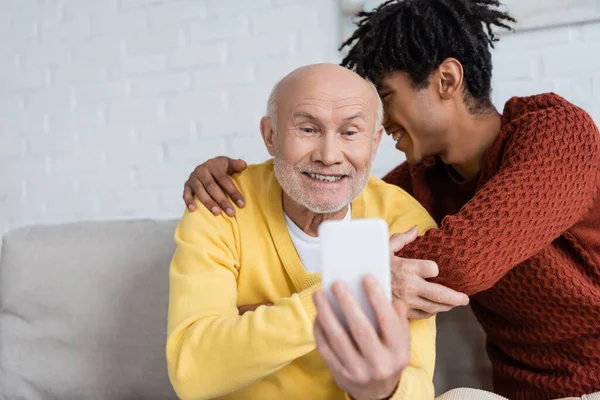 Smiling african american grandson hugging grandparent with smartphone at home — Stockfoto